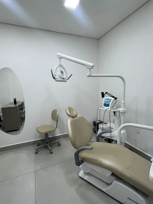 Consultório branco com cadeira odontológica bege e espelho redondo