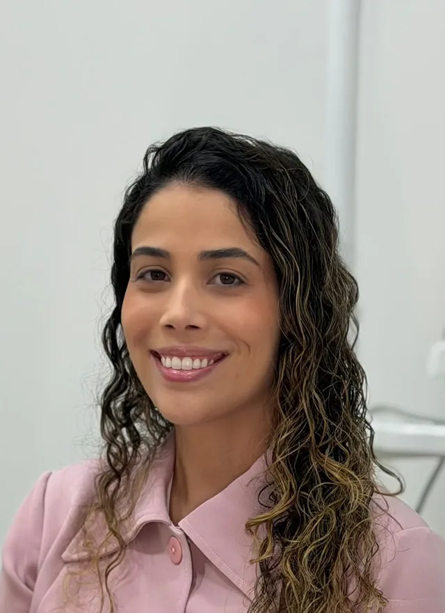 Dra. Yasmin Côrtes Freitas sorrindo, de camisa rosa, no consultório